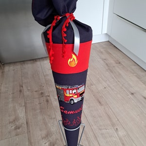Könnte beinhalten: Ein roter und schwarzer Schultüte mit einem Feuerwehrauto-Design und dem Namen "Samuel" bestickt. Die Tüte steht auf einem Metallständer.