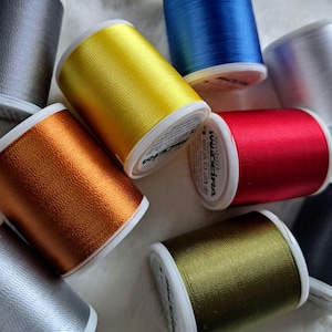 Könnte beinhalten: Sortiment von Spulen mit Stickgarn in verschiedenen Farben, darunter Gold, Silber, Rot, Blau, Grün und Orange. Die Spulen sind mit "Madeira" und "100% Polyester" beschriftet.