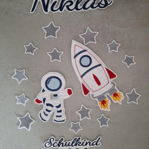 Könnte beinhalten: Ein Set aus gestickten Stoffaufnähern mit einer weißen Rakete mit roten und blauen Details, einem Astronauten im weißen Anzug und mehreren grauen Sternen. Der Text lautet oben Niklas und unten Schulkind 2026 in dunkelblauer Schreibschrift.