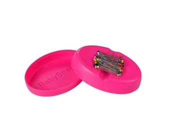 May include: Pink plastic pin cushion with a magnetic lid. The cushion has a slot for holding pins and the lid has the text "BabySnap" printed on it.