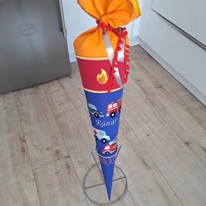 Könnte beinhalten: Ein blauer und orangefarbener Schultüte mit einem Feuerwehrauto- und Autodesign. Die Tüte ist mit einem roten Band und Bommeln verziert. Der Name "Runar" ist auf die Tüte gestickt.