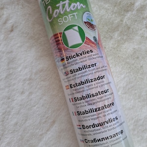 Könnte beinhalten: Eine weiße Rolle abreißbarer Baumwoll-Stabilisator für Stickerei. Das Etikett lautet "Tear Away Cotton Soft" und listet den Produktnamen in mehreren Sprachen auf: Stickvlies, Stabilizer, Estabilizador, Stabilisateur, Stabilizzatore, Borduurvlies und Стабилизатор. Das Etikett enthält auch die Größe: 50 cm x 10 m (19,7" x 11 yds) und die Artikelnummer: Art. 9412.
