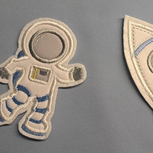 Könnte beinhalten: Zwei gestickte Bügelbilder, eines mit einem Astronauten in einem weißen Raumanzug mit blauen Akzenten und das andere mit einer Rakete mit blauen und weißen Akzenten.