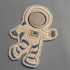 Könnte beinhalten: Gestickter Aufnäher mit einem weißen Astronauten im Raumanzug mit blauen und grauen Akzenten. Der Aufnäher hat eine strukturierte Optik und ist mit detaillierten Nähten versehen, ideal für Kleidung oder Accessoires.