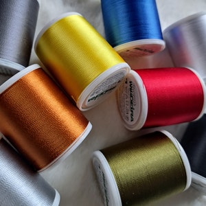 Könnte beinhalten: Sortiment von Garnrollen in verschiedenen Farben, darunter Gold, Rot, Grün, Blau und Silber. Die Rollen sind kreisförmig auf einer weißen Oberfläche angeordnet.