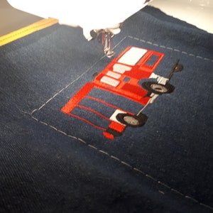 May include: A red and black embroidered design of a fire truck on a blue fabric. The design is being stitched on a sewing machine.