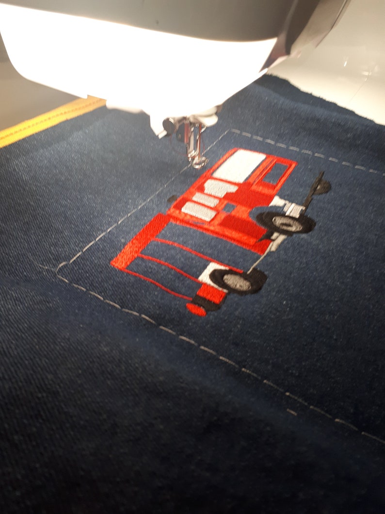 May include: A red and black embroidered design of a fire truck on a blue fabric. The design is being stitched on a sewing machine.