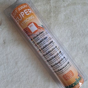 Könnte beinhalten: Eine Rolle weißer, superstarker, weggeschnittener Stabilisator für Stickerei. Die Verpackung zeigt den Produktnamen "Cut Away Super Strong" und listet die Verwendung des Produkts in mehreren Sprachen auf.