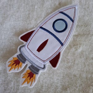 Könnte beinhalten: Gestickter weißer Raketenaufnäher mit roten, blauen und gelben Akzenten. Der Aufnäher zeigt ein detailliertes Design einer Rakete mit Flammen und einem Bullauge. Ideal für Kleidung oder Accessoires.