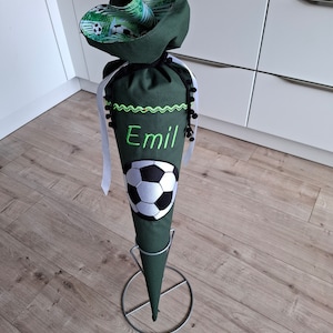 May include: A green fabric cone with a white soccer ball design and the name "Emil" embroidered on it. The cone is on a metal stand.