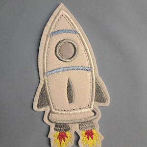 Könnte beinhalten: Eine weiße Rakete mit blauen Streifen und einer grauen Spitze. Die Rakete hat rote und gelbe Flammen, die aus dem Boden kommen.