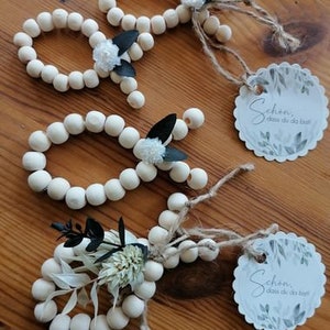 Könnte beinhalten: Drei Holzperlen-Girlanden mit weißen Blüten und schwarzen Blättern. Jede Girlande hat ein Schild mit dem Text "Schön, dass du da bist!" in schwarzer Schrift.