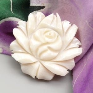 Peut inclure: Une broche en forme de rose, de couleur blanc cassé. La broche est finement sculptée, avec des pétales détaillés pour imiter une fleur épanouie. La broche est posée sur un tissu à motifs verts et violets.