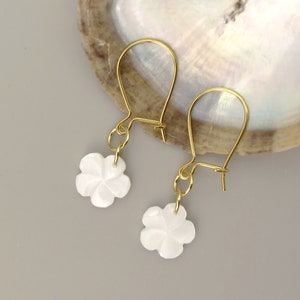 Könnte beinhalten: Ein Paar goldene Ohrringe mit weißen, blumenförmigen Anhängern. Die Ohrringe hängen an einer weißen Oberfläche.