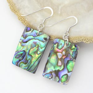 Könnte beinhalten: Ein Paar rechteckige Ohrringe mit schillernden Abalone-Muscheln. Die Ohrringe haben ein silbernes Haken-Design. Die Abalone-Muscheln zeigen eine Mischung aus grünen, blauen und violetten Farbtönen. Die Ohrringe werden auf einer weißen Oberfläche präsentiert.