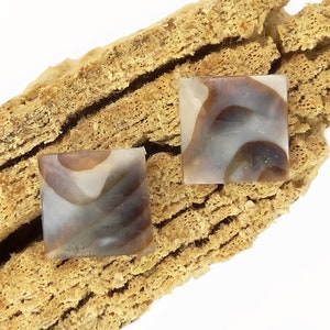 May include: Two square-shaped, brown and white agate cabochons with a swirling pattern. The cabochons are on a piece of driftwood.
