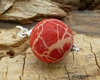 May include: A round, red pendant with a coral-like texture and a silver bail. The pendant has a light beige, web-like pattern across its surface. The jewelry is displayed on a weathered wooden surface.