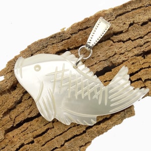 Könnte beinhalten: Ein perlmuttfarbener Fischanhänger mit geätzten Details, der an einer silberfarbenen Öse befestigt ist. Das Fischdesign weist einen detaillierten Schwanz und Körpermarkierungen auf. Der Anhänger wird vor einem strukturierten, braunen Hintergrund präsentiert.