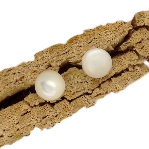 Puede incluir: Un par de pendientes de botón de perlas blancas redondas con un acabado liso y brillante. Las dos perlas circulares están colocadas una al lado de la otra, descansando dentro de la grieta natural, texturizada y dentada de un trozo de corteza marrón claro.