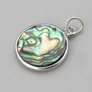 May include: A round pendant featuring a polished abalone shell inlay with iridescent green, blue, and purple swirls, encased in a smooth silver-toned metal frame with a small circular jump ring attached to the top for hanging on a chain.