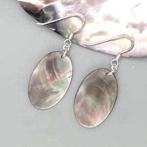 Könnte beinhalten: Ein Paar Ohrhänger mit ovalen, irisierenden Anhängern aus dunkelgrauer Abalone-Muschel. Die Anhänger hängen an silberfarbenen Metallhaken und zeigen natürliche, wirbelnde Muster in violetten, grünen und rosafarbenen Farbtönen.