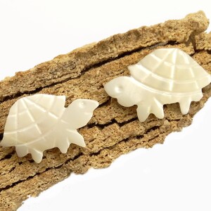 Könnte beinhalten: Zwei weiße Perlmutt-Anhänger in Form von Schildkröten. Die Schildkröten sind mit einem detaillierten Panzermuster geschnitzt und haben eine glatte, polierte Oberfläche.