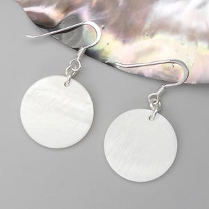May include: A pair of white, circular earrings with a pearlescent sheen. The earrings are suspended from silver-colored fish hook ear wires. The background is a light gray surface with a portion of a pearlescent shell.
