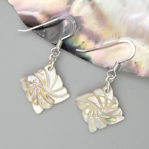 Könnte beinhalten: Ein Paar Ohrhänger mit floralem Design. Die Ohrringe haben einen perlmuttartigen, fächerförmigen Anhänger mit geschnitzten Details, der an silberfarbenen Fischhaken-Ohrdrähten hängt. Die Ohrringe sind vor einem grauen Hintergrund platziert.