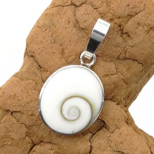 Könnte beinhalten: Ein silberner Anhänger mit einem weißen, oval geformten Muscheldesign. Die Muschel hat ein Spiralmuster in Weiß- und Cremetönen. Der Anhänger hat eine silberne Öse zum Anbringen an einer Kette. Der Hintergrund ist eine strukturierte, braune Oberfläche.