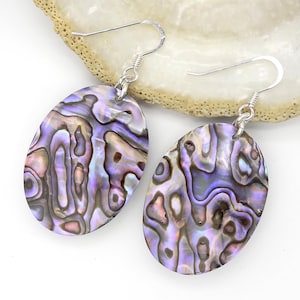 Könnte beinhalten: Ein Paar oval geformte Abalone-Muschel-Ohrringe mit irisierenden Wirbeln in Violett, Blau und Rosa. Die polierten Muschelstücke hängen an silberfarbenen Metallhaken und weisen eine natürliche, mehrfarbige und glänzende Oberfläche auf.