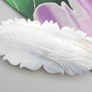 Könnte beinhalten: Ein weißer, geschnitzter Haarclip aus Perlmutt in Blütenform. Der Clip hat die Form einer Blume mit detaillierten Blütenblatt- und Blattmustern. Der Hintergrund umfasst eine graue Oberfläche und einen Stoff mit grünen und lila floralen Designs.