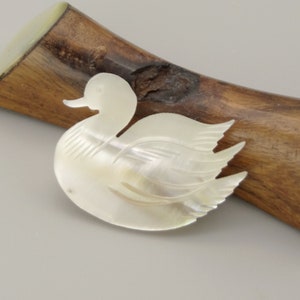 Op de afbeelding: Een witte parelmoer gegraveerde eenden broche. De eend is in een zwemmende positie met zijn kop naar de zijkant gedraaid. De broche is op een houten standaard.