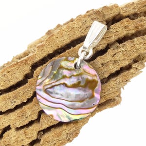 Könnte beinhalten: Ein runder Anhänger mit schillerndem Abalone-Muscheleinsatz, der Farbtöne von Rosa, Grün und Gold aufweist. Der Anhänger hat eine silberne Öse zum Anbringen an einer Kette. Der Anhänger wird auf einem Stück strukturiertem, hellbraunem Holz gezeigt.