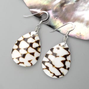 May include: A pair of teardrop earrings with a white and brown pattern. The earrings have silver-colored hooks. The earrings are displayed on a gray surface with a pearlescent background.