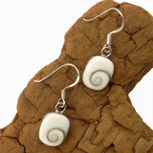 May include: Silver earrings with a white, rectangular pendant and a spiral design. The earrings have a hook fastening and are displayed on a piece of brown wood. These earrings are a jewellery item.
