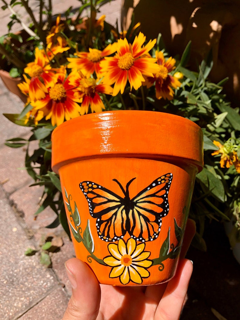 May include: Orange terracotta flower pot with a hand-painted monarch butterfly and a yellow flower design.