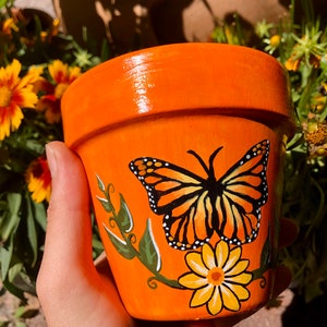 May include: Orange terracotta flower pot with a hand-painted monarch butterfly and yellow flower design.