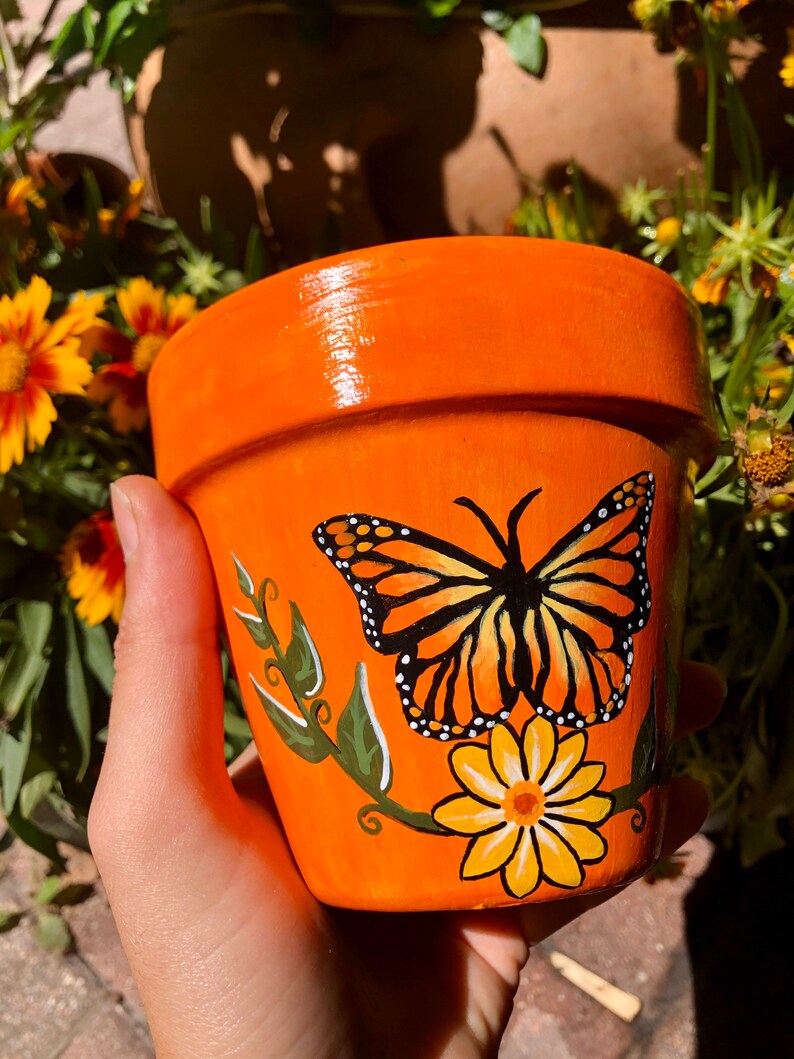 May include: Orange terracotta flower pot with a hand-painted monarch butterfly and yellow flower design.