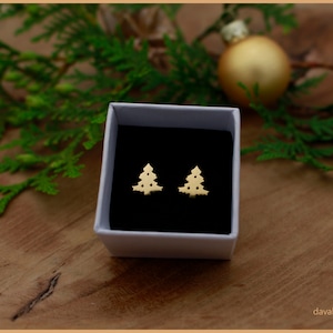 Könnte beinhalten: Ein Paar goldfarbene Weihnachtsbaum-Ohrringe, präsentiert in einer weißen Schachtel mit schwarzem Innenraum. Die Ohrringe haben die Form stilisierter Weihnachtsbäume. Der Hintergrund umfasst Grün und eine goldene Kugel.