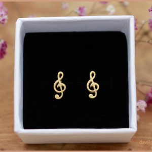 Könnte beinhalten: Ein Paar goldfarbene Musiknoten-Ohrringe in einer weißen Schachtel mit schwarzem Innenraum. Die Ohrringe sind in Form von Violinschlüsseln.