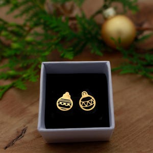 Könnte beinhalten: Ein Paar goldfarbene Ohrstecker in Form von Weihnachtskugeln, präsentiert in einer weißen Schachtel mit schwarzem Innenraum. Die Ohrringe haben ein festliches Design, vor dem Hintergrund von Grün und einer goldenen Kugel.