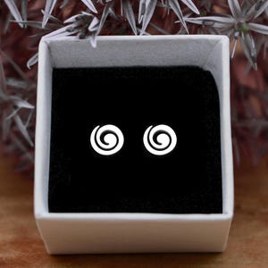Könnte beinhalten: Ein Paar weiße Spiral-Ohrstecker in einer weißen Schmuckschatulle mit schwarzem Samtinnenraum. Die Ohrringe sind klein und kreisförmig, mit einem minimalistischen Design. Die Schatulle ist geöffnet und zeigt die Ohrringe.
