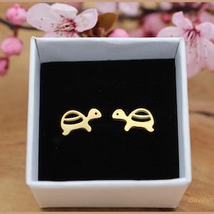 Könnte beinhalten: Ein Paar goldfarbene Schildkröten-Ohrstecker in einer weißen Schachtel mit schwarzem Innenraum. Die Ohrringe sind klein und zart und perfekt, um jedem Outfit einen Hauch von Verspieltheit zu verleihen.