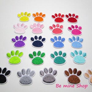 Könnte beinhalten: Eine Sammlung bunter Pfotenabdruck-Patches in verschiedenen Farben, darunter Gelb, Orange, Rot, Rosa, Lila, Blau, Grün und Braun. Jeder Patch hat einen weißen Rand und ist gestickt.