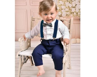 May include: A baby boy wearing a white button-down shirt with a plaid bow tie and navy blue suspenders. He is also wearing navy blue pants and is sitting on a white wooden chair.