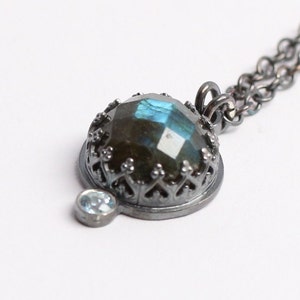 Könnte beinhalten: Eine silberne Halskette mit einem schwarzen Metallanhänger, der einen großen blauen Labradorit-Edelstein zeigt, der von einem dekorativen Filigranmuster umgeben ist. Ein kleinerer, klarer Edelstein ist am Anhänger befestigt.