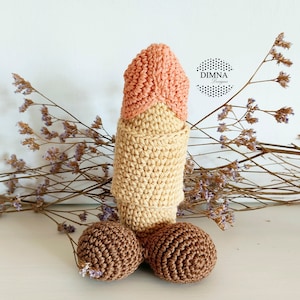 May include: A crocheted plush toy shaped like a human male anatomy. The toy is made with beige, brown, and peach colored yarn. The toy is sitting on a white surface with dried flowers in the background.