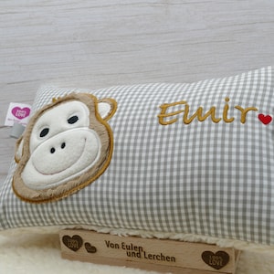 May include: A rectangular pillow with a gray and white checkered pattern. The pillow features an embroidered monkey face in shades of brown and white. The name "Emir" is embroidered in gold thread, with a small red heart. A wooden stamp is visible.