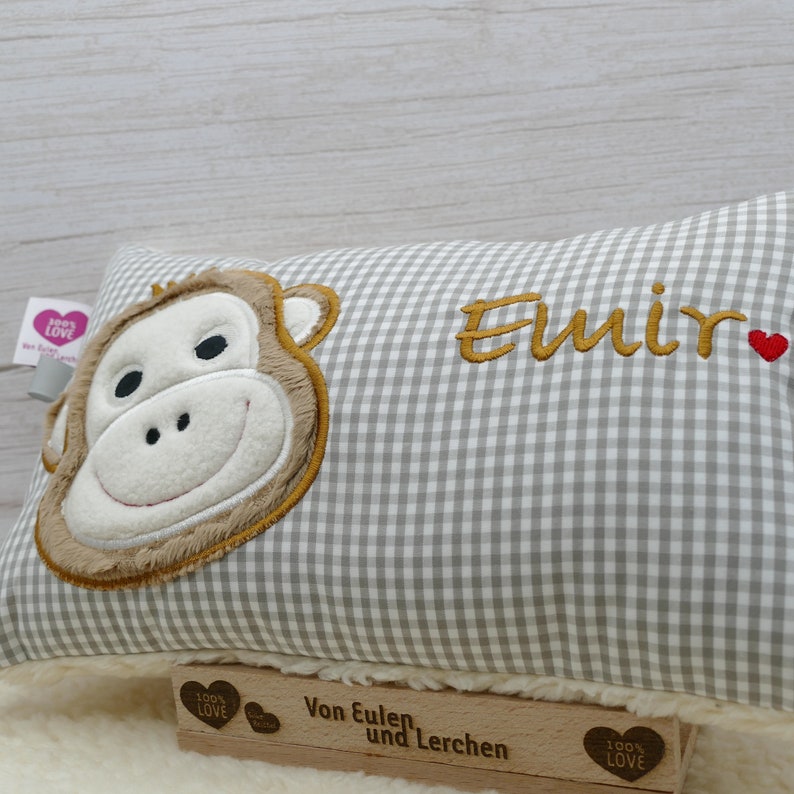 May include: A rectangular pillow with a gray and white checkered pattern. The pillow features an embroidered monkey face in shades of brown and white. The name "Emir" is embroidered in gold thread, with a small red heart. A wooden stamp is visible.