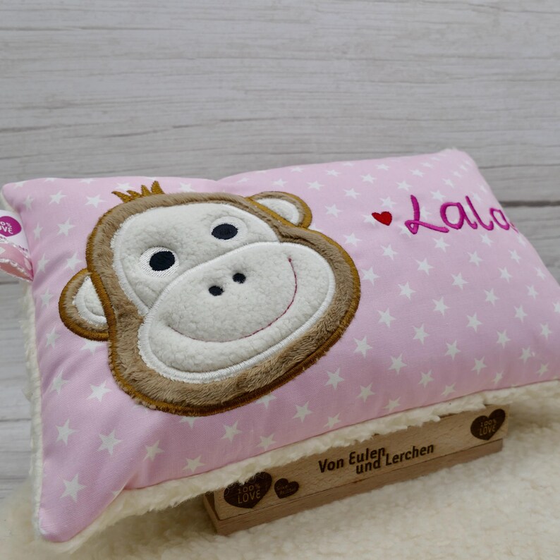 May include: Pink rectangular pillow with a monkey face applique. The monkey has a white face, brown ears and a smiling mouth. The pillow has white stars and the name "Lalo" embroidered on it. The pillow is resting on a white fluffy surface.